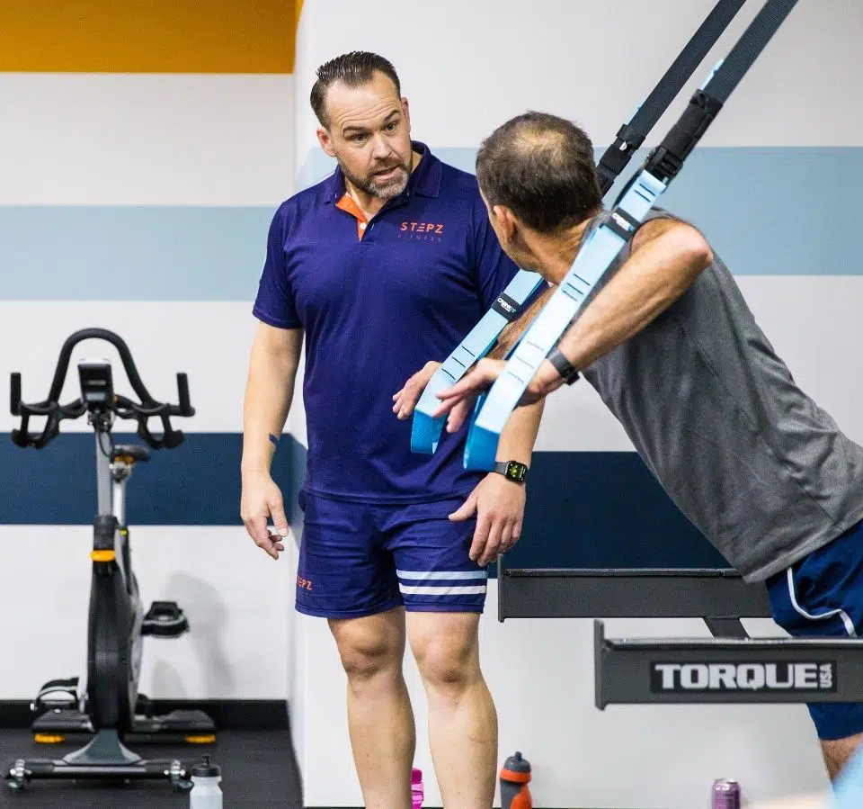 Instructor guiding a person using suspension training gear in a 24/7 gym.