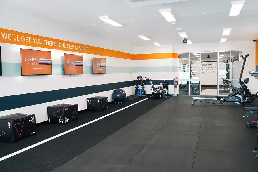 Modern gym with exercise equipment, plyometric boxes, wall-mounted TVs, and motivational signage.