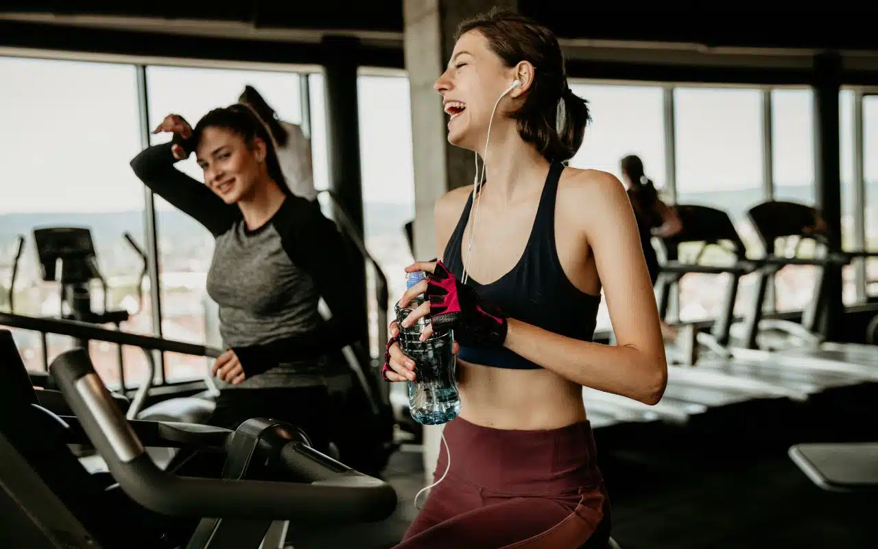 Two women at a gym on cardio machines, one laughing—enjoying no lock-in contracts.