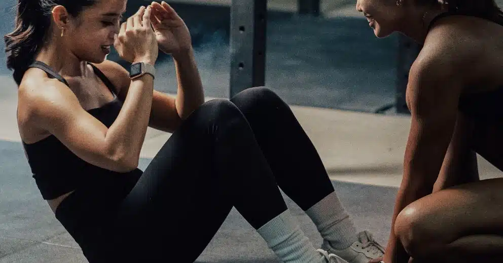 Two women show ways to make time for the gym as one assists the other with sit-ups.